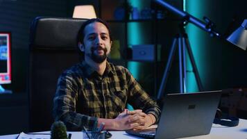 Portrait of happy man sitting at home office desk, preparing to solve tasks on laptop. Cheerful freelancer in front of digital device, doing email communication in living room, camera A video