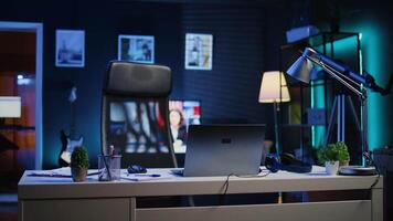 Focus on office desk in front of smart TV in blurry background displaying news program broadcasting. Laptop on table and television screen in empty room showing VOD channel, camera B video