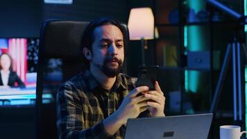 Man relaxing in apartment sitting at office desk, texting friends on cellphone, enjoying leisure time. Smiling remote worker taking break to talk with mates on internet using phone, camera A video