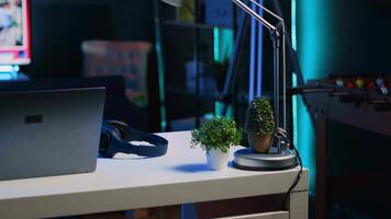 Apartment illuminated by RGB lights with office desk in front of smart TV displaying news program broadcasting. Laptop, mini house plants and headphones on table in empty room, camera A close up shot video