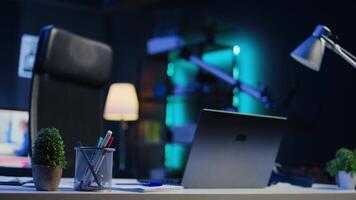 Office desk filled with business productivity items in empty home interior with TV left open in background at night. Laptop, headphones, plants and work documents on table in apartment room, camera B video