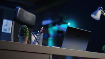 Office desk filled with productivity items in empty home interior illuminated with neon lights at night. Laptop on table and other work related objects in apartment room, camera B close up shot video