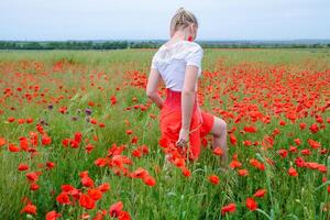 Blonde young woman in red skirt and white shirt, red earrings is in the middle of a poppy field. photo