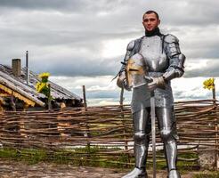 Knight in armor on the background of a wicker fence, a wooden house and the expanses of the motherland. photo