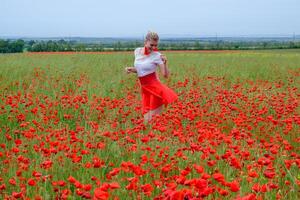 Blonde young woman in red skirt and white shirt, red earrings is in the middle of a poppy field. photo