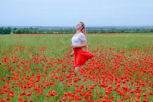 Blonde young woman in red skirt and white shirt, red earrings is in the middle of a poppy field. photo