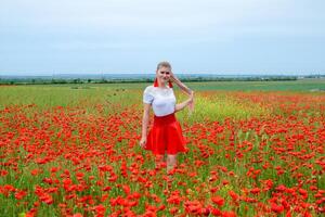 Blonde young woman in red skirt and white shirt, red earrings is in the middle of a poppy field. photo