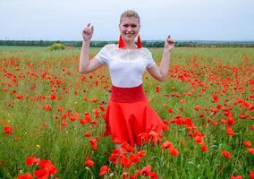 Blonde young woman in red skirt and white shirt, red earrings is in the middle of a poppy field. photo