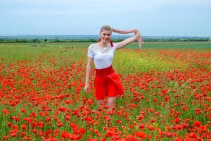 Blonde young woman in red skirt and white shirt, red earrings is in the middle of a poppy field. photo