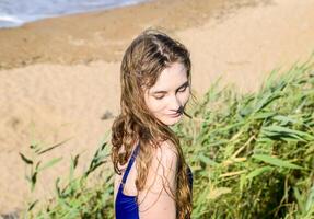 Beautiful blonde in a blue bathing suit on a background of green reeds and the sea. The girl looks down. photo