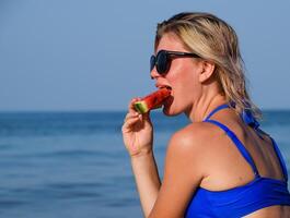 A blonde in sunglasses eats a watermelon by the sea. A juicy watermelon in the hands of a woman photo