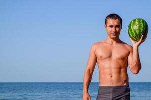 A sporty man is standing on the beach and holding a whole watermelon. A ripe watermelon in the hands of a man photo