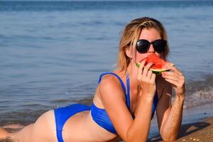 The blonde in sunglasses lies on the beach near the waves and eats a watermelon. A juicy watermelon in the hands of a woman. photo