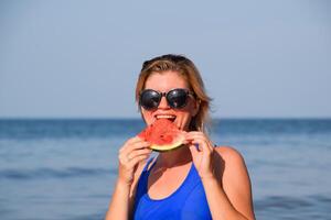 A blonde in sunglasses eats a watermelon by the sea. A juicy watermelon in the hands of a woman photo