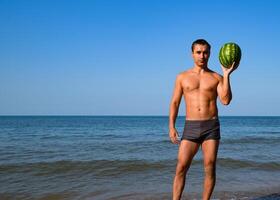 A sporty man is standing on the beach and holding a whole watermelon. A ripe watermelon in the hands of a man photo