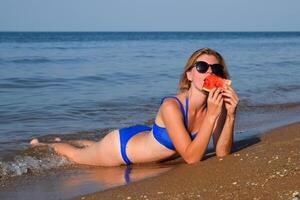 The blonde in sunglasses lies on the beach near the waves and eats a watermelon. A juicy watermelon in the hands of a woman. photo