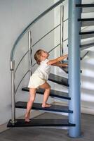 pequeño niño niña yendo arriba escalera a moderno hogar, niño alpinismo espiral escalera. peligroso situación a hogar. niño la seguridad concepto. foto