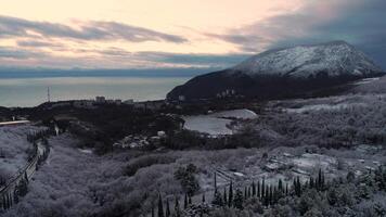 panorámico ver de el terraplén de el ciudad y nevadas alto montaña en puesta de sol. disparo. aéreo para un costero ciudad, invierno arboles y campos, alto montaña cubierto con nieve en contra azul mar. video