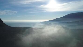 Aerial view of endless ocean, coastline near high mountain in clouds. Shot. Stunning view of a rock in a shadow of the sun, blue ocean, small town down in the valley on cloudy sky background. video