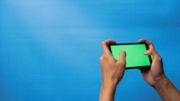 Hand of man holding mobile smartphone green screen with game playing gesture on blue background photo