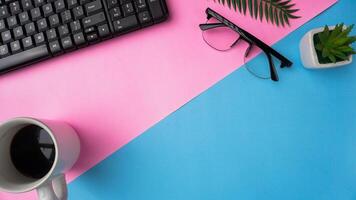 workspace desk with keyboard, glasses, plants, and coffee on blue and pink background photo