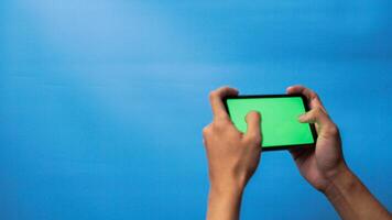 Hand of man holding mobile smartphone green screen with game playing gesture on blue background photo