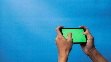 Hand of man holding mobile smartphone green screen with game playing gesture on blue background photo