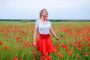 Blonde young woman in red skirt and white shirt, red earrings is in the middle of a poppy field. photo