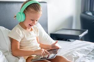 Happy little girl in headphones using digital tablet and smiling happy while listening to music or playing game in home bed. High quality photo