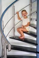 pequeño niña es sentado solo en espiral escalera a moderno hogar. peligroso situación a hogar. niño la seguridad concepto. foto