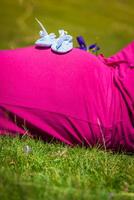 Pregnant woman lying on a grass with baby shoes on her belly photo