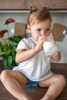 contento bebé niña sentado a el mesa en el cocina y Bebiendo Leche foto