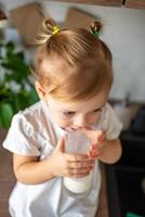 contento bebé niña sentado a el mesa en el cocina y Bebiendo Leche foto