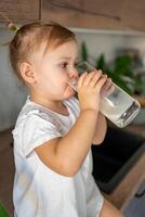 Happy baby girl sitting at the table in the kitchen and drinking milk photo