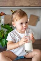 contento bebé niña sentado a el mesa en el cocina y Bebiendo Leche foto