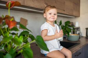 Happy baby girl sitting at the table in the kitchen and drinking milk photo