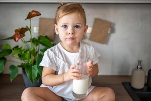 Happy baby girl sitting at the table in the kitchen and drinking milk photo