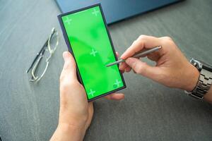 Close up view of man hand holding smart phone with green chroma key screen and stylus pen at his creative workspace photo