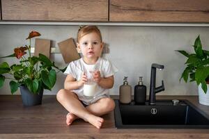 Happy baby girl sitting at the table in the kitchen and drinking milk photo