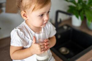 Happy baby girl sitting at the table in the kitchen and drinking milk photo