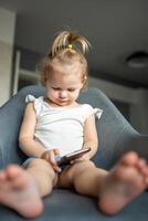 Smiling little girl is sitting on armchair and using smart phone photo