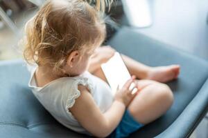 Smiling little girl is sitting on armchair and using smart phone photo