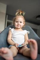 Smiling little girl is sitting on armchair and using smart phone photo