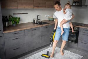 Father with his daughter on his hands vaccuming the floor in the modern home kitchen photo