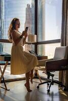 Stylish businesswoman freelancer working with smart phone and sitting on table, enjoying panoramic view in city background. Low key photo. High quality photo