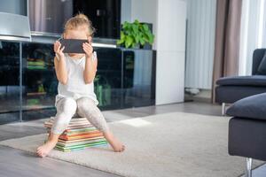 Cute Little girl sits on stack of children's books and watches cartoons on smartphone photo
