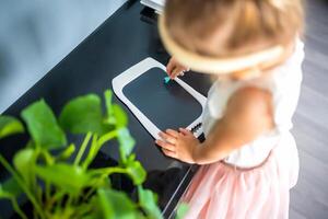 pequeño niña es dibujo en LED tableta a hogar. ver de niño mano con pintura Escribiendo foto