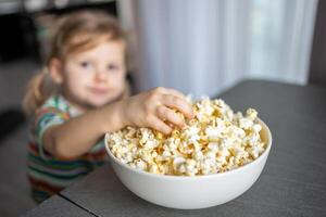 pequeño niña es comiendo palomitas de maiz en hogar cocina. atención en mano tomando palomitas de maiz foto