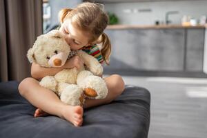 Cute little girl hugging teddy bear while sitting on sofa at home photo