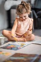 pequeño niña se sienta en el piso a hogar y sorteos con pinturas y cepillos en un colorante libro. temprano infancia creatividad y educación. alto calidad foto